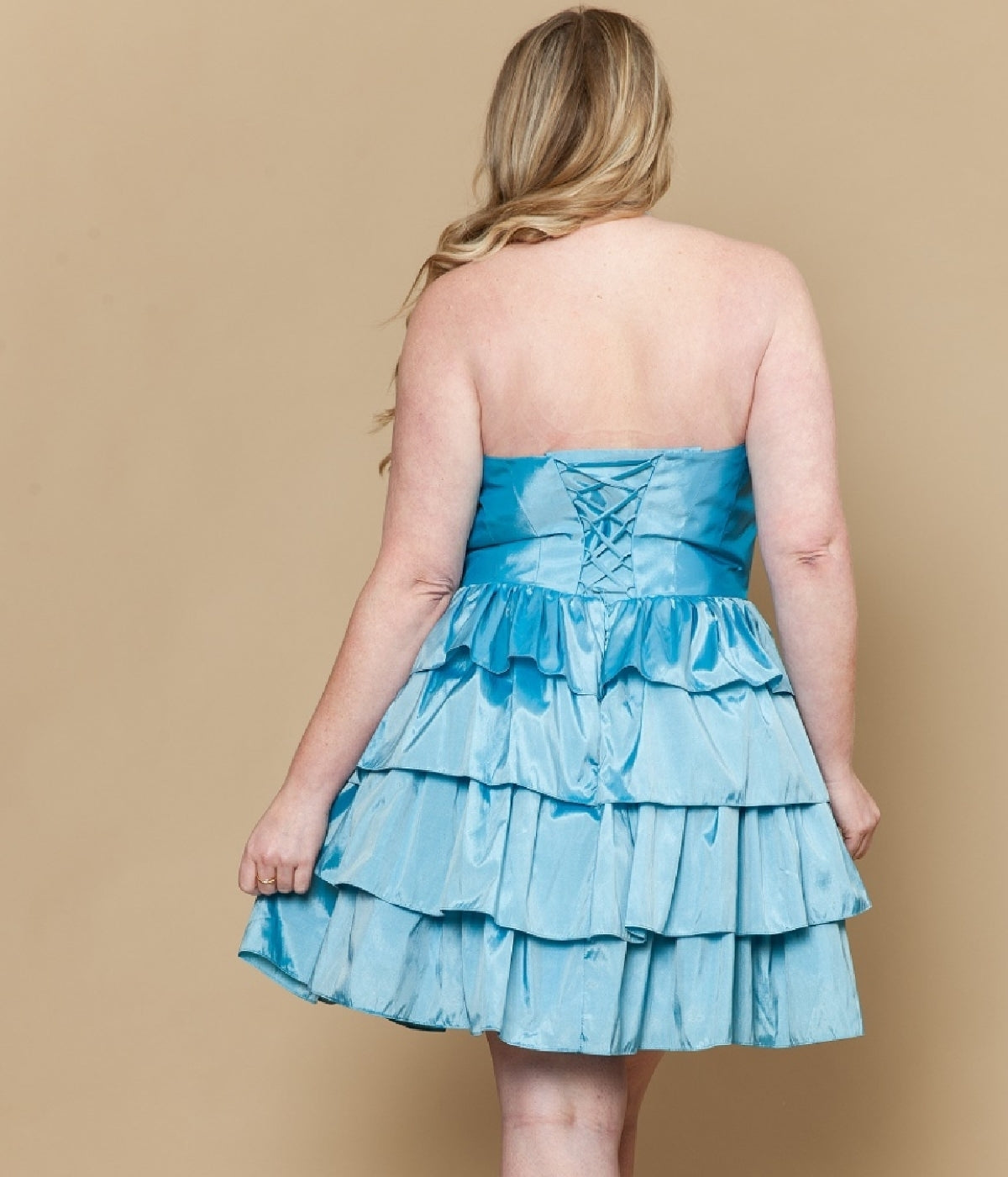 A woman with long blonde hair is seen from the back wearing the Poly USA W1216 Plus-Size Short Halter Party Dress, featuring blue ruffled tiers and a lace-up bodice, perfect for semi-formal occasions, against a beige background.